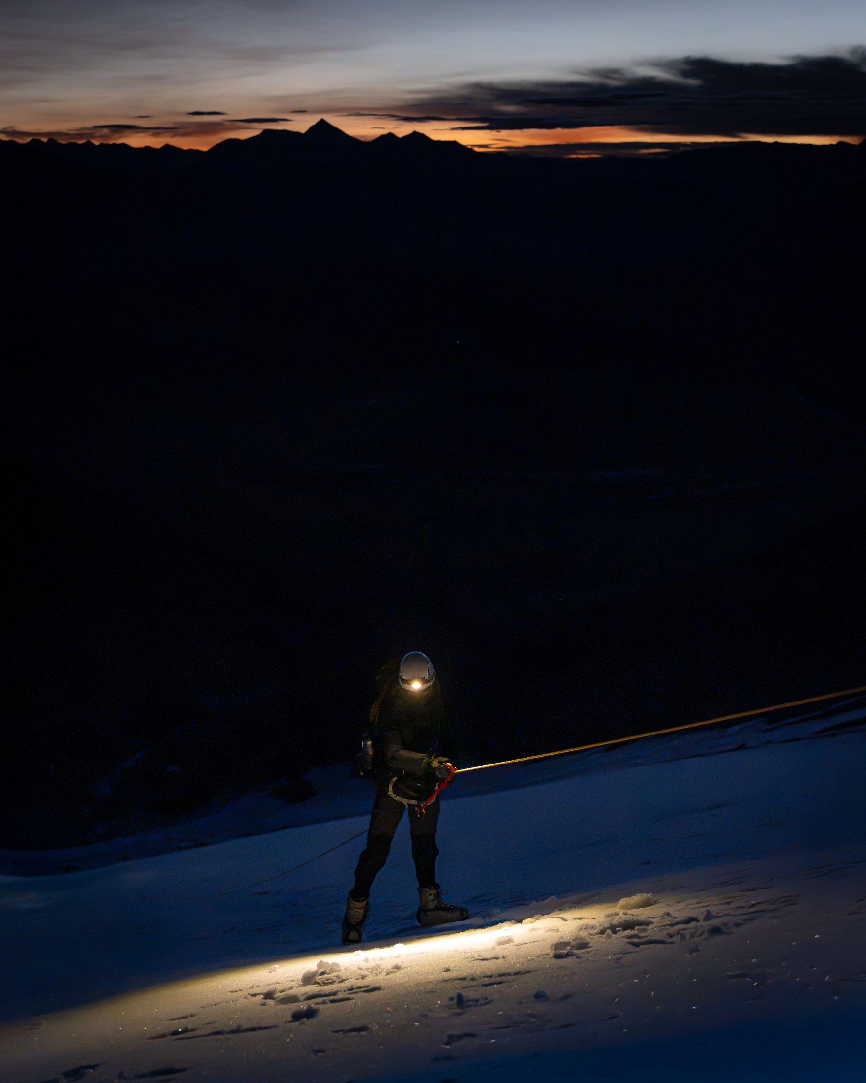 A lone climber lights the snow with a headlamp beneath a twilight horizon